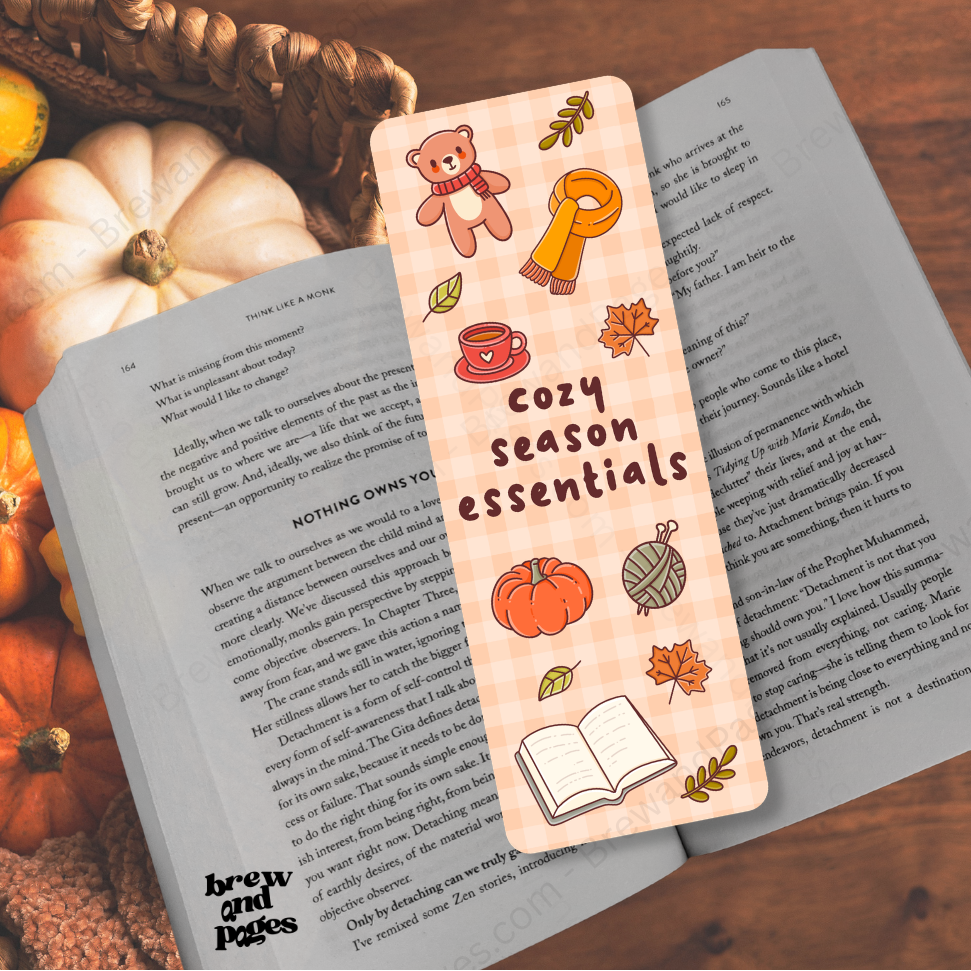 A rectangular paper bookmark with a peach plaid pattern and autumn-themed illustrations, including a teddy bear, pumpkin, coffee cup, key, yarn ball, leaves, and an open book. The bookmark is placed over an open book on a wooden surface, with pumpkins and a basket in the background.