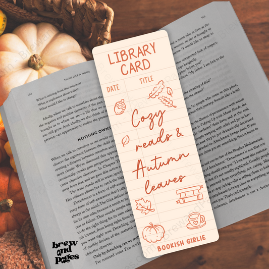 A cream-colored library card bookmark with orange illustrations and text reading 'Library Card' and 'Cozy reads & Autumn leaves' by Bookish Girlie, placed over an open book on a wooden surface with pumpkins and a basket in the background.