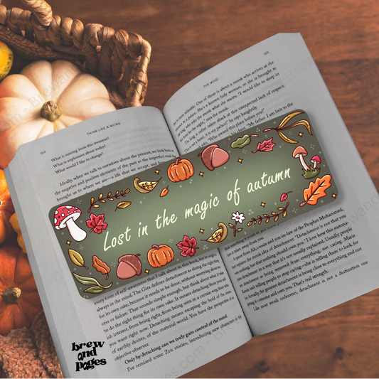 An open book on a wooden surface with autumn-themed bookmarks. One bookmark is placed over the left page and reads 'Lost in the magic of autumn' with illustrations of pumpkins, acorns, leaves, and mushrooms.